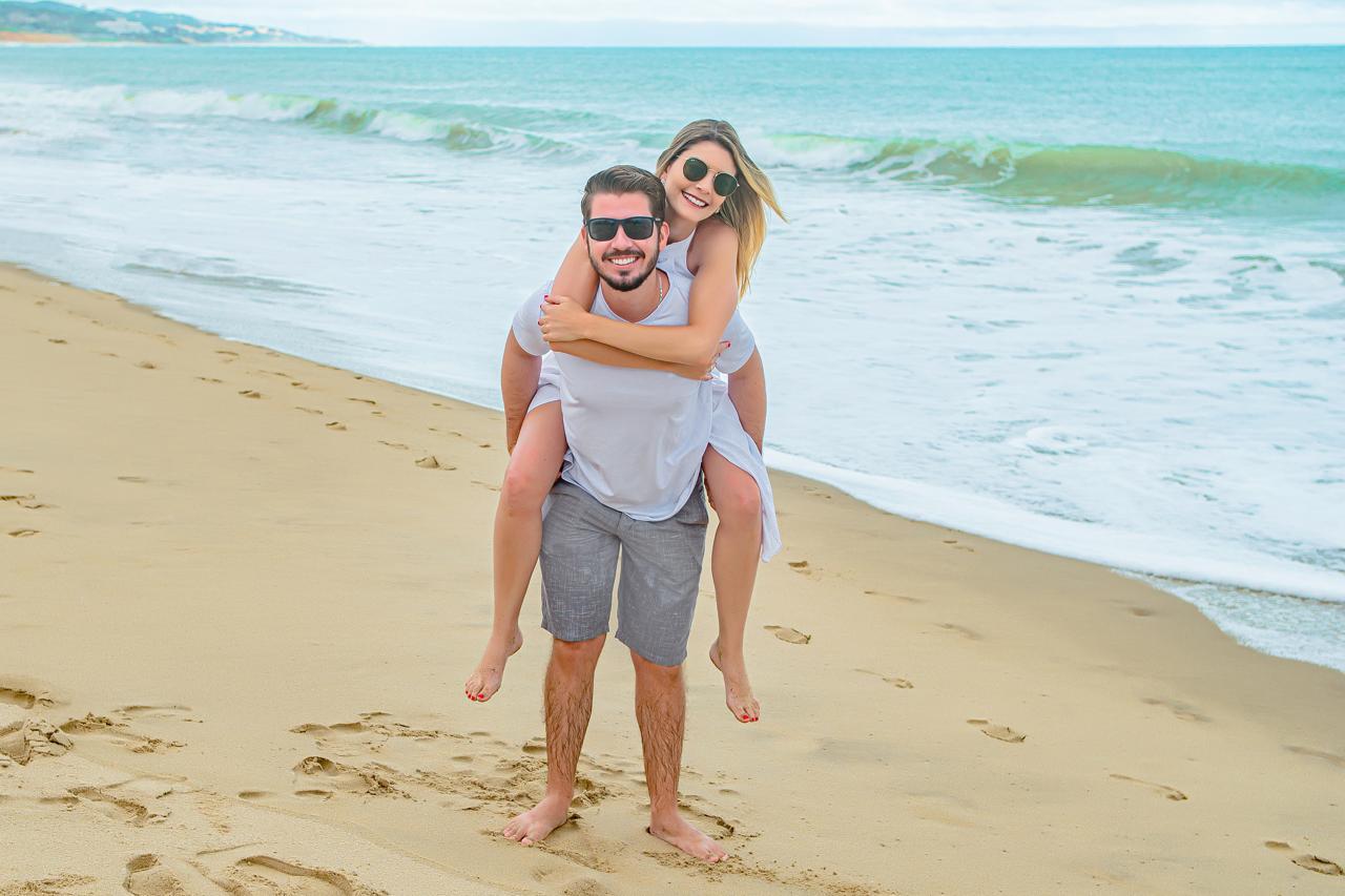 Gustavo carregando Caroline nas costas, sorrindo na praia